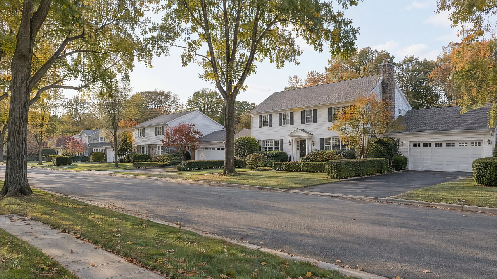 A tree-lined street of white colonial houses with attached two-car garages, clipped hedges and asphalt driveways in early autumn