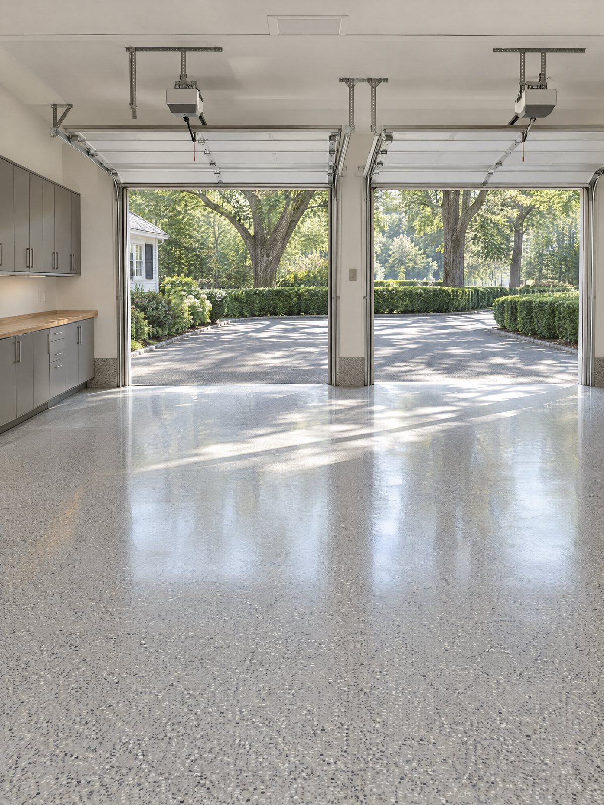A two-car garage with a gray and white epoxy flake floor, both overhead doors open to a driveway edged with clipped hedges and oak trees, gray cabinets and an oak workbench along one wall